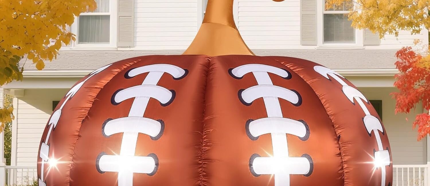 Football-themed Halloween inflatable pumpkin with LED lights glowing in a front yard surrounded by colorful fall leaves.