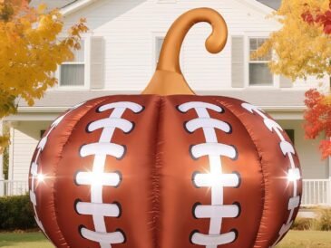 Football-themed Halloween inflatable pumpkin with LED lights glowing in a front yard surrounded by colorful fall leaves.