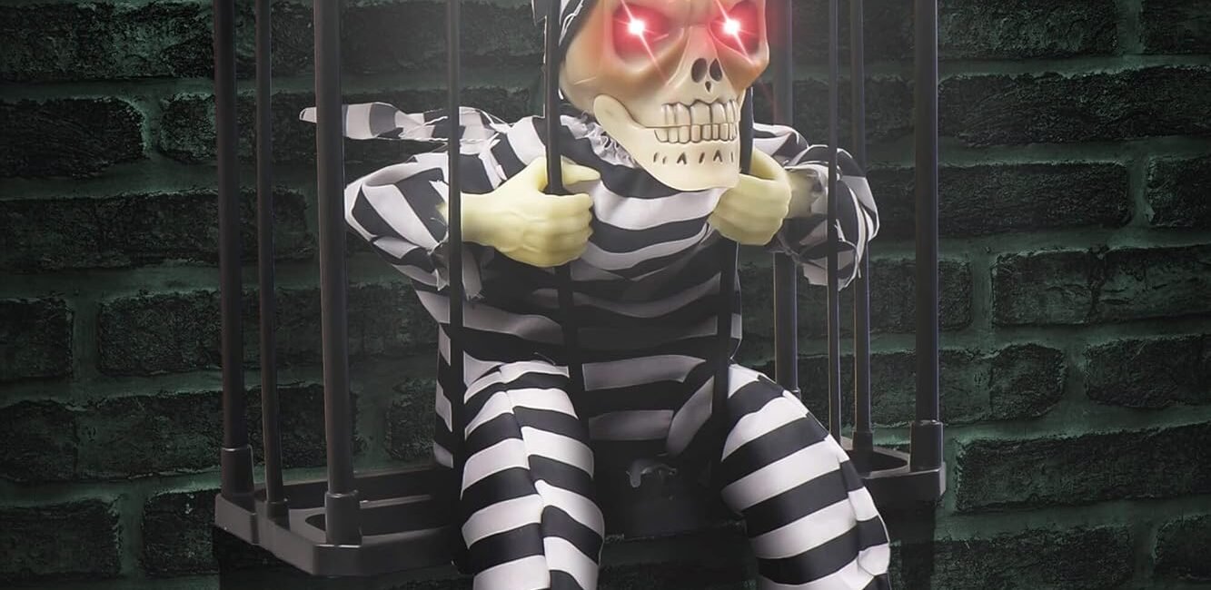 Halloween skeleton prisoner decoration in a cage with glowing red eyes, motion sensor, and striped jumpsuit hanging against a dark brick wall.