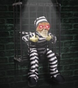 Halloween skeleton prisoner decoration in a cage with glowing red eyes, motion sensor, and striped jumpsuit hanging against a dark brick wall.