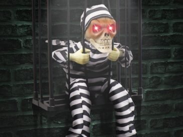 Halloween skeleton prisoner decoration in a cage with glowing red eyes, motion sensor, and striped jumpsuit hanging against a dark brick wall.