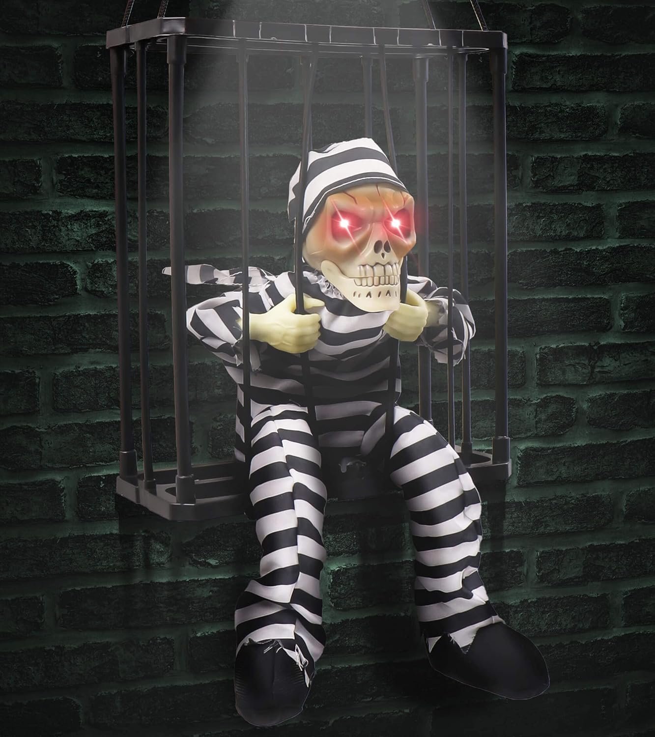Halloween skeleton prisoner decoration in a cage with glowing red eyes, motion sensor, and striped jumpsuit hanging against a dark brick wall.