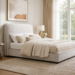 Pacifico upholstered bed with light neutral fabric, curved headboard, warm brown trim, cream bedding, and a textured rug in a bright modern bedroom.
